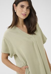 Rövid ujjú, V-nyakú ruha világoszöld anyagból, puha textúrával, oldalsó zsebekkel és diszkrét varrásokkal a formázás érdekében.