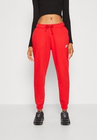 Červené tepláky s elastickým pasem a šnůrkou, zdobené malým bílým logem Nike. V kombinaci s černými sportovními botami.