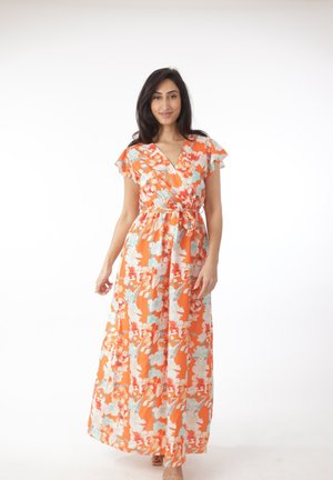 Mujer de pie vistiendo un vestido largo naranja con flores, sin mangas y con un lazo en la cintura, sobre un fondo blanco liso.