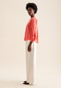 Koraliowa bluzka z haftem kwiatowym na rękawach, zestawiona z kremowymi spodniami o szerokich nogawkach. Modelka nosi czarne szpilki. Miękka, teksturowana tkanina.