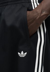 Χέρι στην τσέπη μαύρου παντελονιού Adidas με λευκές τρεις ρίγες και λογότυπο Adidas trefoil στο μηρό.