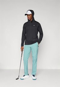 Fekete cipzáras kapucnis pulóver kapucnival, világoskék golfnadrág, fehér golfcipő. Golfütőt tart, fehér sapkát visel.