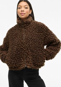 Smeđa pahuljasta jakna s visokim ovratnikom i prednjim zatvaračem, koja se ističe teksturiranom površinom i voluminoznom siluetom, u kombinaciji s crnim hlačama.