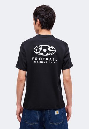 Personne portant un t-shirt noir avec un logo de football blanc et le texte « Football Training Gear » dans le dos, associé à un jean bleu.