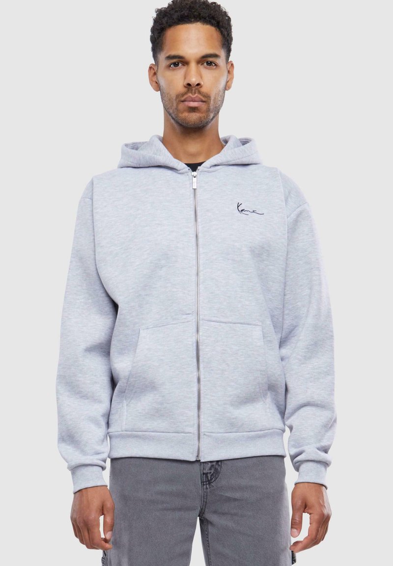 Light gray zip-up hoodie made of soft fabric, featuring a kangaroo pocket, ribbed cuffs, and a small black logo embroidered on the chest.
