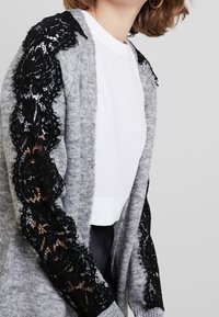 Grijze cardigan met lange mouwen en zwarte kanten details op de schouders en zijkanten. Draagt over een wit shirt, met een zachte textuur.