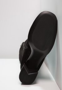 Botín de ante negro con suela de goma texturizada y tacón triangular. Presenta un acabado suave y detalles de diseño minimalistas.