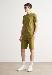 Set de tricou din bumbac de culoare verde măslin și pantaloni scurți cu detalii ale logo-ului, asortat cu adidași albi și șosete albe, stând în fața unui fundal deschis.