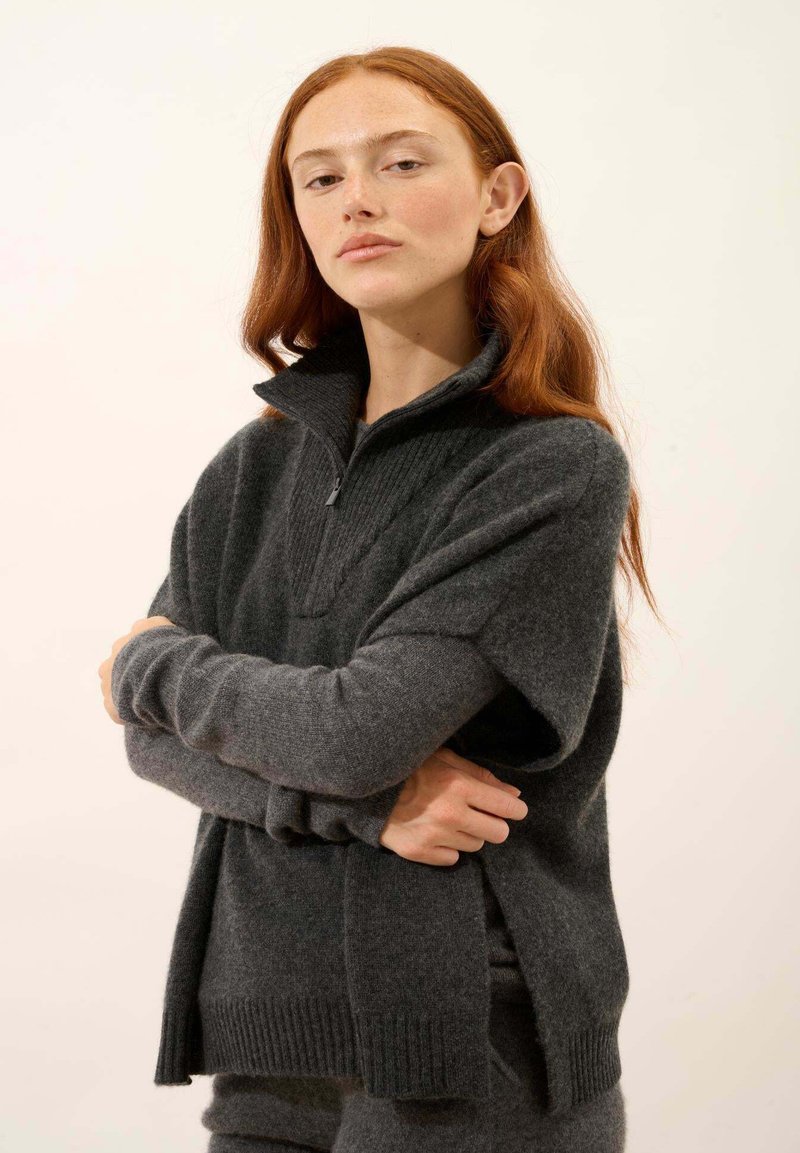 Pullover grigio a maglia con colletto alto, texture a coste e spacchi laterali. Il tessuto appare morbido e pesante, con maniche lunghe e ampie.