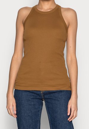 Kobieta ubrana w brązową, bezrękawnikową, prążkowaną koszulkę, połączoną z ciemnoniebieskimi dżinsami na jednolitym tle.