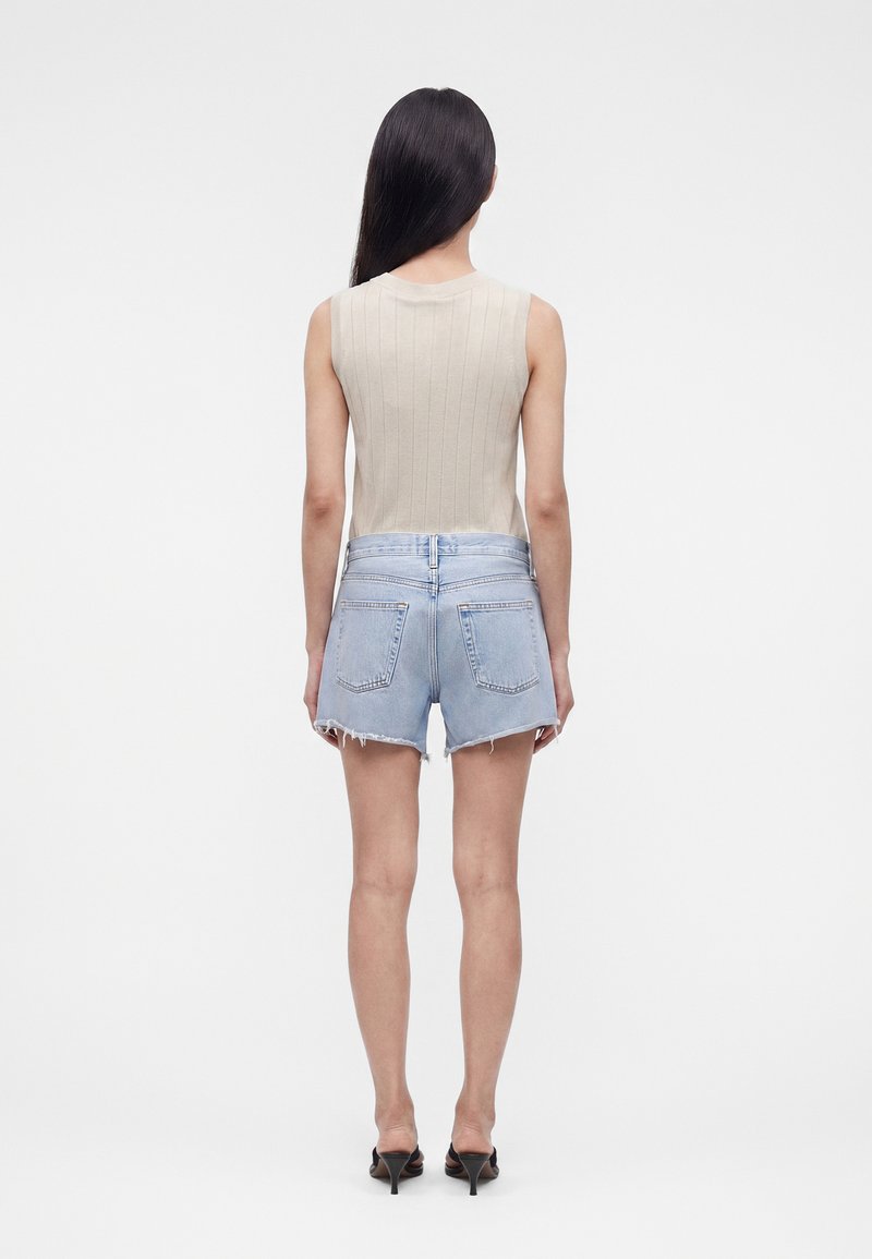 Femme aux longs cheveux noirs portant un haut côtelé beige sans manches, un short en jean bleu clair avec ourlets bruts et des sandales à talons noires, vue de dos.