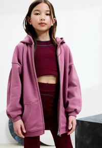 Prevelký zipsový sveter v svetloružovej farbe, s prednou kapsou, rebrovanými manžetami a mäkkou textúrou, nosený cez vínový crop top.