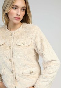 Veste en fausse fourrure crème avec un col rond, fermeture à boutons et deux poches sur la poitrine décoratives avec des boutons brillants. Texture douce et design ajusté.