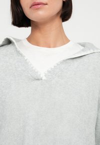 Ανοιχτό γκρι φούτερ με κουκούλα και απαλή υφή, με V-λαιμόκοψη και λευκή λεπτομέρεια κατά μήκος της κουκούλας, φορεμένο πάνω από ένα λευκό T-shirt.