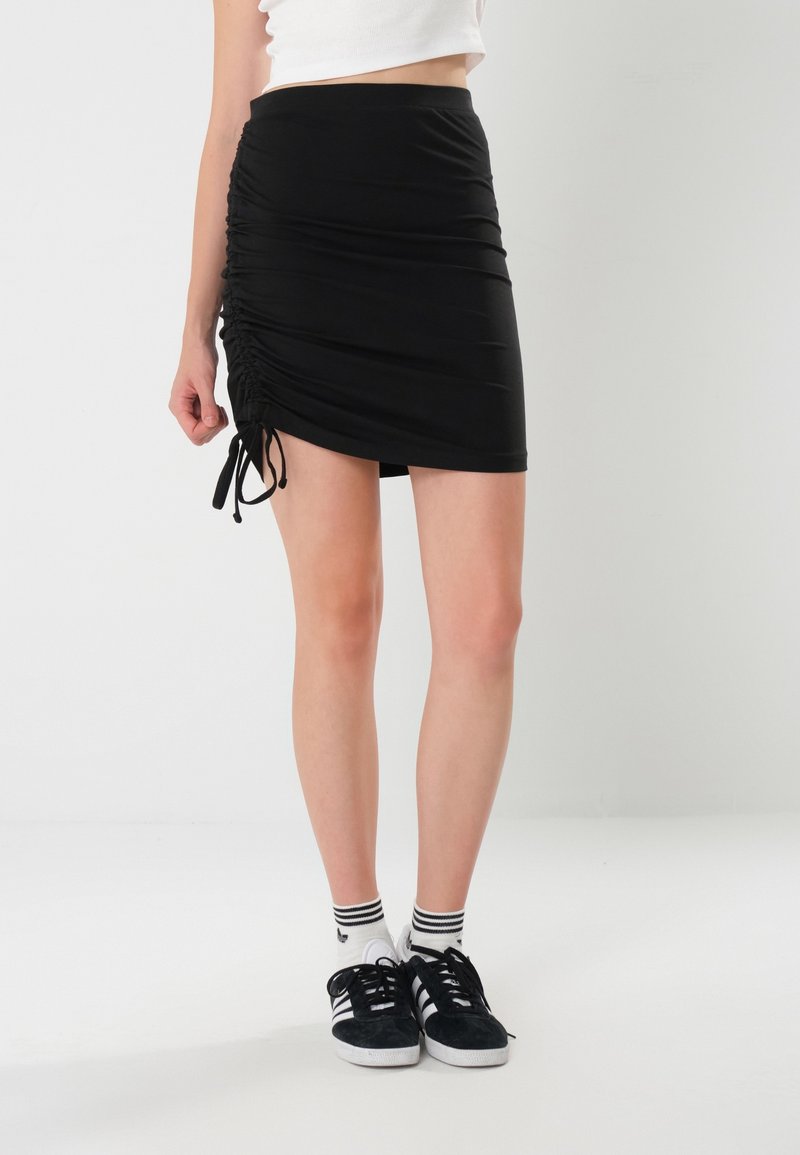 Mini-jupe noire en tissu extensible avec des détails froncés sur les côtés et des liens ajustables. Assortie à des baskets noires et des chaussettes rayées.
