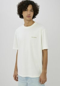 T-shirt en coton blanc à coupe décontractée, avec des manches courtes et un texte vert brodé discret sur le côté gauche de la poitrine.