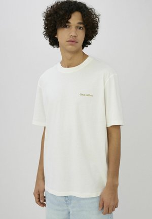 T-shirt en coton blanc à coupe décontractée, avec des manches courtes et un texte vert brodé discret sur le côté gauche de la poitrine.