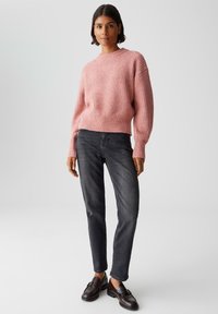 Frau steht in einem rosa gestrickten Pullover, dunkelschwarzen Jeans und dunkelbrown Leder-Slippern vor einem schlichten hellen Hintergrund.