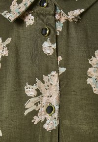 Olivenfarvet skjorte med beige blomstermønster og sorte knapper med guldkanter, der viser detaljer ved krave og knaplukning.