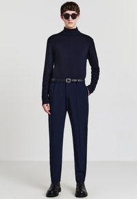 Navy-Turtleneck-Pullover kombiniert mit maßgeschneiderten navyfarbenen Hosen. Ein schwarzer Gürtel, elegante schwarze Stiefel und dunkle Sonnenbrillen runden den Look ab.