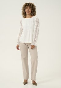 White lace blouse with long sleeves, diamond pattern, round neckline. Paired with beige trousers and green heels.