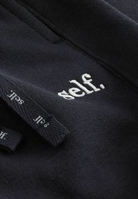 Svart tyg med broderat "self." i vitt. Har texturerad sömnad och varumärkesetiketter, vilket framhäver en minimalistisk design.