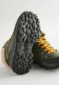 Buty trekkingowe z ciemnozieloną tkaniną, czarną gumową podeszwą, wytrzymałym wzorem bieżnika oraz żółtymi i czarnymi sznurówkami, z puszystym różowym akcentem.
