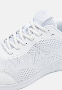 Chaussure de sport blanche avec une tige en mesh perforée, lacets plats et semelle en caoutchouc. Accent logo sur le côté et la languette.