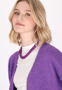 Cárdigan de punto púrpura con escote en V, usado sobre una blusa blanca. Presenta un collar de cadena gruesa púrpura, textura suave y un aspecto acogedor.
