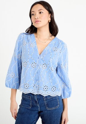 Frau trägt eine hellblaue, bestickte Bluse mit floralen Ausschnitten und 3/4-Ärmeln, kombiniert mit dunkelblauen Jeans.