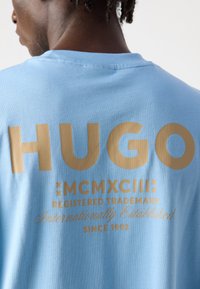 Svijetloplava pamučna majica sadrži podebljani zlatni tekst: "HUGO," "MCMXCIII," "Registrirani zaštitni znak," i "Međunarodno osnovana od 1993."