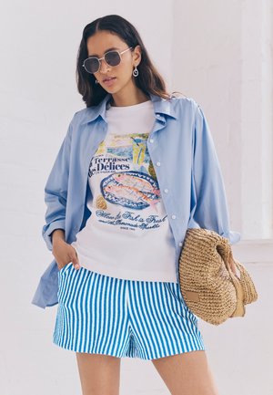 Ljusblå knäppskjorta över en vit t-shirt med grafisk tryck som har fiskdesign, tillsammans med blå och vita randiga shorts och en vävd väska.