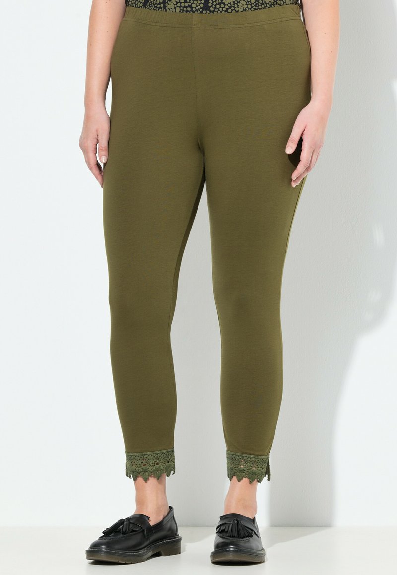 Femme portant un pantalon ajusté vert olive avec de la dentelle sur les chevilles et des mocassins à franges noirs, se tenant contre un fond blanc.