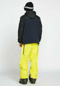 Schnee-Outfit in Schwarz und Gelb, bestehend aus einer schwarzen, isolierten Jacke mit Kapuze und leuchtend gelben, wasserdichten Hosen, ergänzt durch schwarze Stiefel.