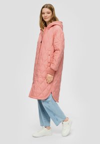Gepolsterter rosa langer Mantel mit Kapuze, ausgestattet mit einem Reißverschluss und Druckknöpfen, kombiniert mit hellblauen weiten Jeans und weißen Sneakers.