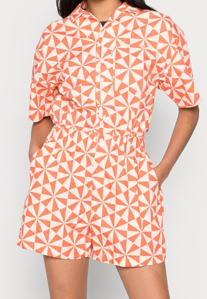Kraťasový overal s oranžovým a bílým geometrickým vzorem, který má zapínání na knoflíky, elastický pas a boční kapsy.