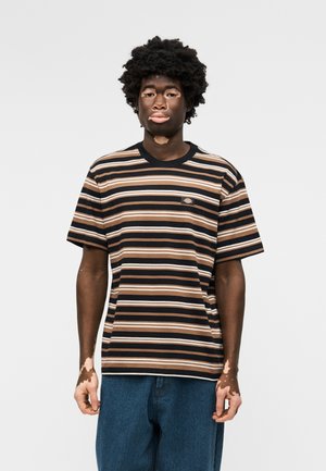 Νεαρό άτομο με λεύκη που φοράει μαύρη, καφέ και άσπρη ριγέ μπλούζα και μπλε τζιν, στέκεται μπροστά σε μονόχρωμο γκρι φόντο.