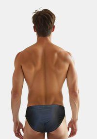 Slips de bain noirs en matériau lisse, présentant un design taille basse, une forme ajustée et une construction minimaliste sans motifs visibles.