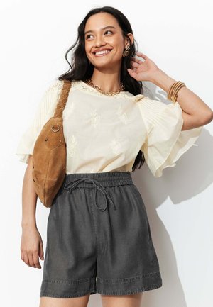 Femme souriante portant un chemisier brodé crème, un short taille haute foncé, un sac à bandoulière tressé marron, des bijoux en or, posant avec la main près des cheveux.