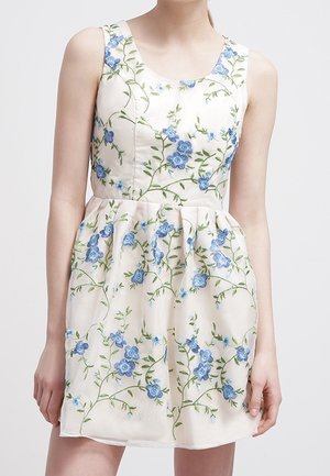 Femme portant une robe crème sans manches avec broderie florale bleue et une taille ajustée sur un fond uni.