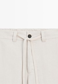 Beige linen pants with elastic waistband, black button, belt loops, and beige fabric drawstring tied at front.