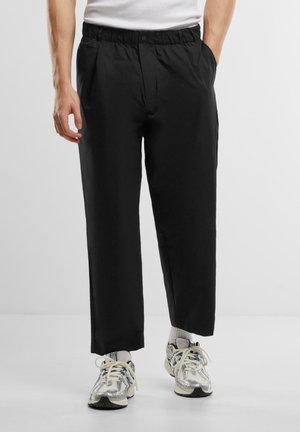 Black trousers with a relaxed fit, featuring an elastic waistband and front pleats. Worn with textured, multi-colored sneakers.