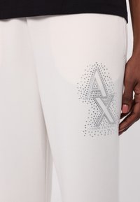 Valkoiset jumppahousut, joissa on kristalleilla koristeltu logo, jossa lukee "AX" ja "Armani Exchange". Silkkinen kangas ja rento malli.