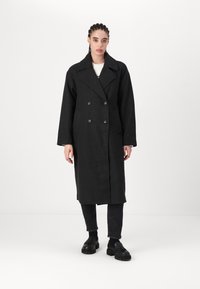 Cappotto in lana nero doppiopetto con ampi rever, tasche frontali e dettagli con bottoni, abbinato a jeans neri e scarpe.
