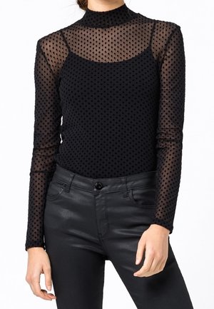 Kobieta ubrana w czarny przezroczysty, w kropki top z długim rękawem nałożony na czarny top, zestawiony z czarnymi skórzanymi spodniami z wysokim stanem.