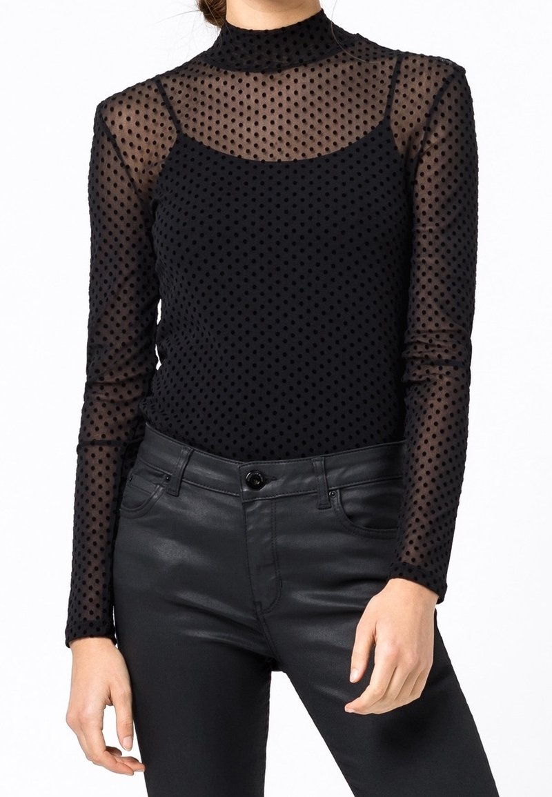 Femme portant un top noir transparent à pois manches longues superposé à un caraco noir, associé à un pantalon en cuir noir taille haute.