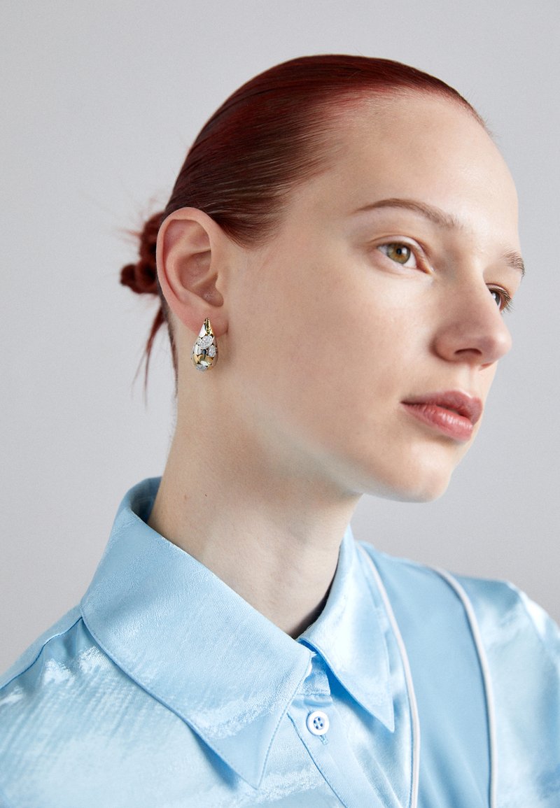 Asara formas pilienu auskari ar zeltītu un caurspīdīgu akmeņu, valkāti uz gaiši ziliem satīna krekla ar strukturētu apkakli un vieglu spīdumu.