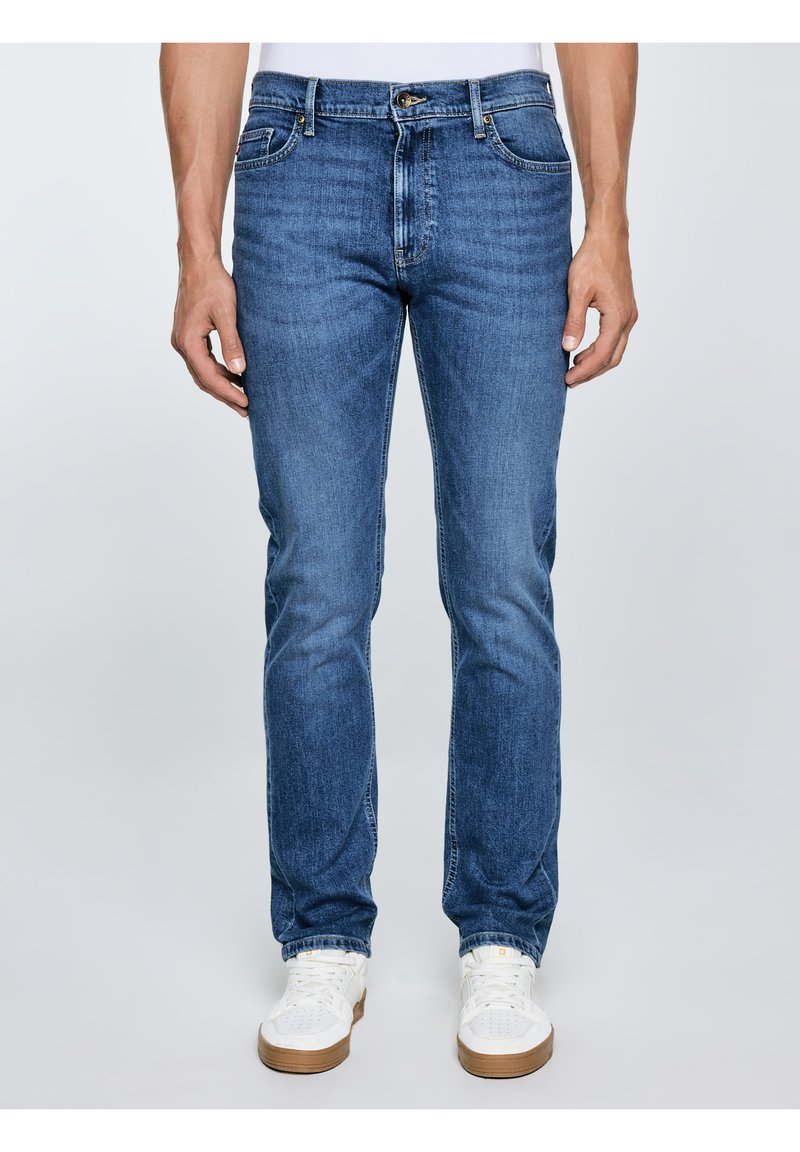 Vīrietis zilās džinsa biksēs un baltos sporta apavos, stāvot pret vienmērīgu gaiši pelēku fonu.