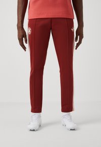 Röda sportbyxor med vita sidostråk, med en logotyp på vänster lår. Tyget verkar slätt och elastiskt. Bärs med vita skor.