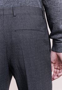Hombre con pantalones de vestir de textura gris oscuro y una camisa de manga larga a juego, mostrando el detalle del bolsillo trasero y con la mano derecha relajada.
